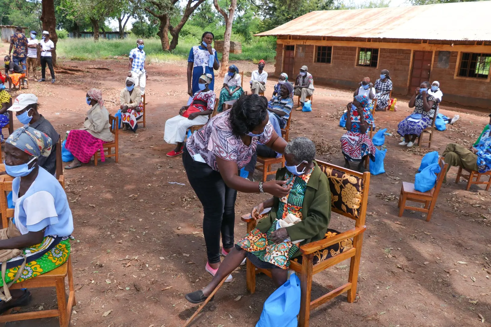 Health workers attend to elderly people seated outdoors in a village clinic yard.