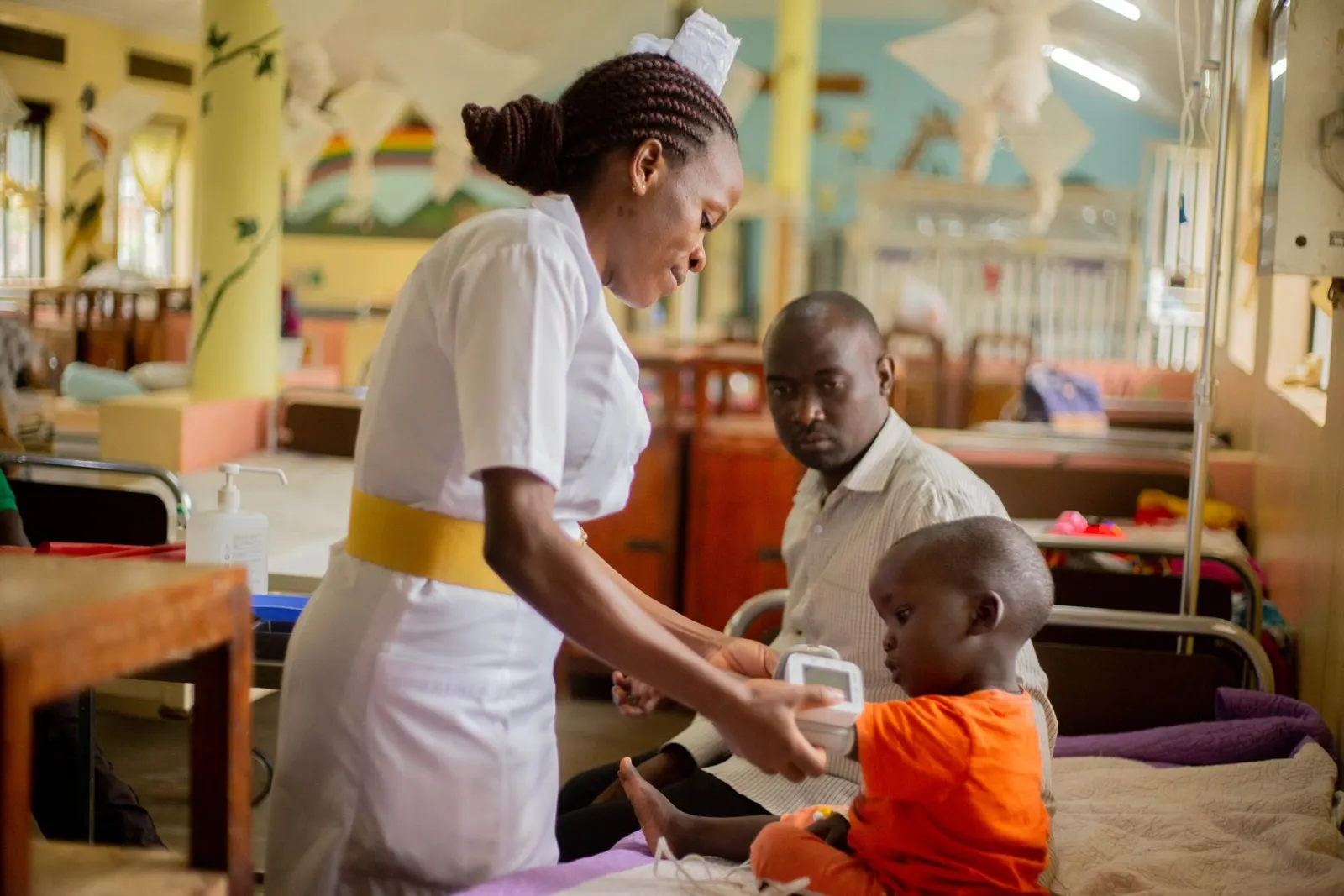 A nurse checks on a young child in a hospital ward while his father looks on.