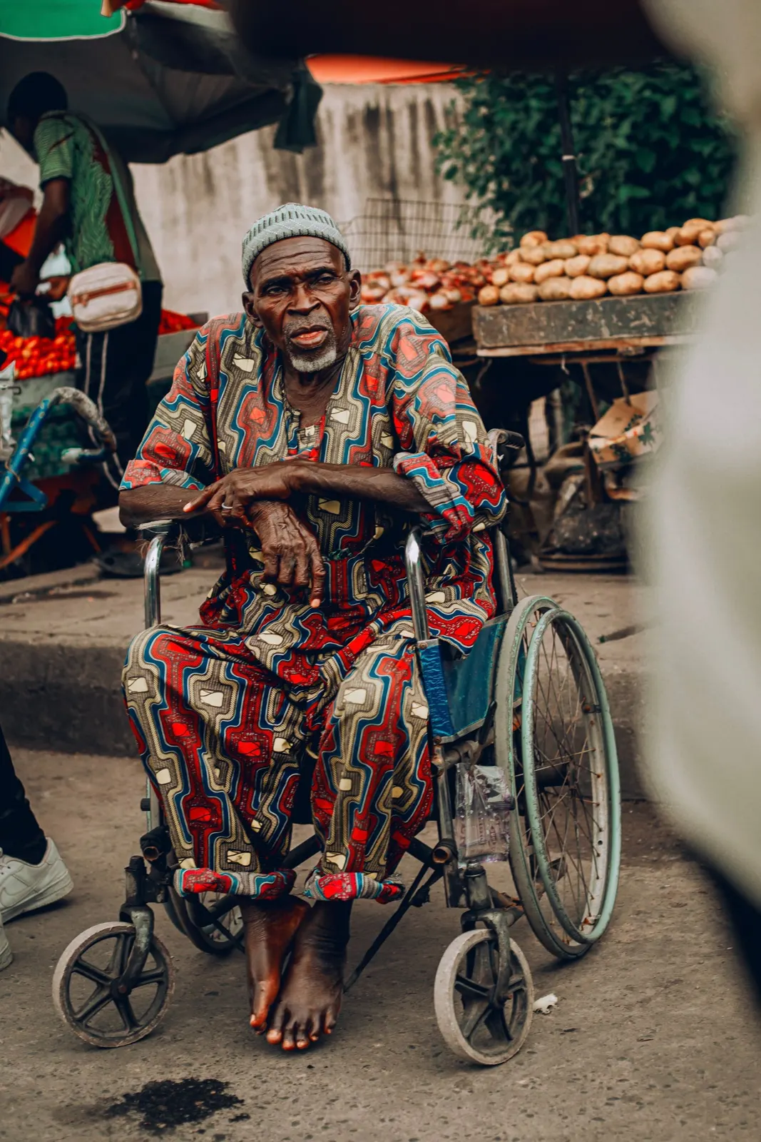An older man in a patterned shirt sits in a wheelchair at a busy street market.