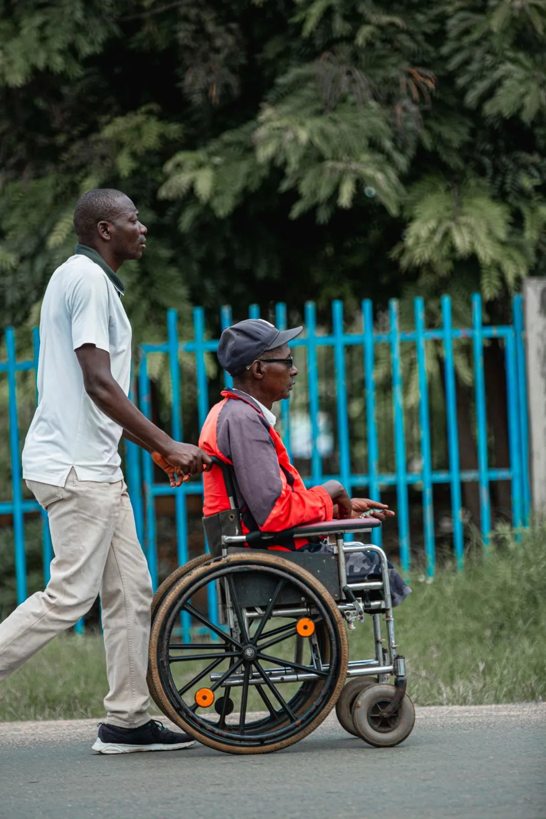 A man pushes a friend in a wheelchair along a quiet street.